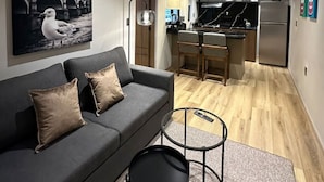 A modern living room with a grey sofa, two black side tables, and a black coffee table. A large black-and-white painting of a bridge and a seagull is on the wall. The room leads to a kitchen with a stainless steel refrigerator, a black countertop, and a hanging light fixture. - Departamento Moderno Soho POP 104 Narvarte (Mexico City)