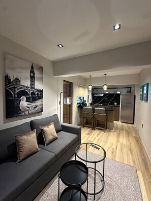 A modern living room with a grey sofa, two black side tables, and a black coffee table. A large black-and-white painting of a bridge and a seagull is on the wall. The room leads to a kitchen with a stainless steel refrigerator, a black countertop, and a hanging light fixture. - Departamento Moderno Soho POP 104 Narvarte (Mexico City)