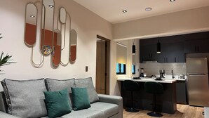 A modern living room with a grey sofa and teal cushions, a kitchen with dark cabinets and a stainless steel refrigerator, and a wall-mounted mirror with a geometric design. - Departamento  Boutique en Narvarte  204 (Mexico City)