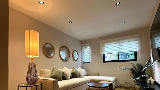 A modern living room with a sofa, a coffee table, and a lamp. There are three wall-mounted mirrors and a window with blinds.