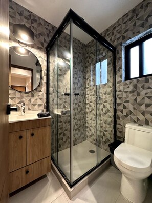A modern bathroom with a glass-enclosed shower, a wooden vanity, a white toilet, and a geometric-patterned wall. - Departamento Moderno y Centrico Estilo Wabi Sabi (Mexico City)