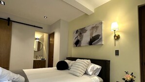 Ein ordentlich bezogenes Bett mit weißen Bettwäsche und einer grauen Tagesdecke, ein Nachttisch mit Blumen in einer Vase und eine Wandlampe.