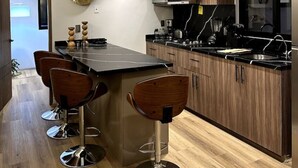 Eine moderne Küche mit Holzausbau, einer schwarzen Arbeitsplatte und Edelstahlgeräten. Drei Barhocker und ein großer dekorativer Teller sind an der Wand zu sehen.
