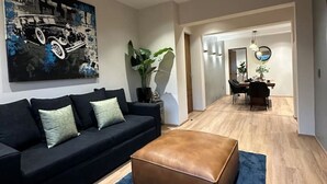 Un salon moderne avec un canapé noir, un pouf marron et un tapis bleu. On aperçoit à l’arrière-plan un coin repas avec une table et des chaises.