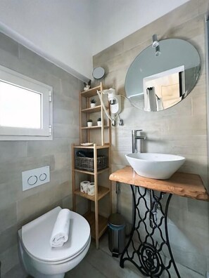 Une salle de bain moderne avec des toilettes blanches, une étagère en bois contenant des articles de toilette, un miroir rond et un lavabo blanc posé sur un plan de travail en bois.