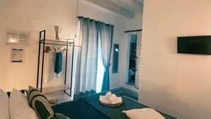A hotel room with a bed, a massage table, a window with curtains, a coat rack, and a wall-mounted TV. - NAP25GUESTHOUSE (Polignano a Mare)