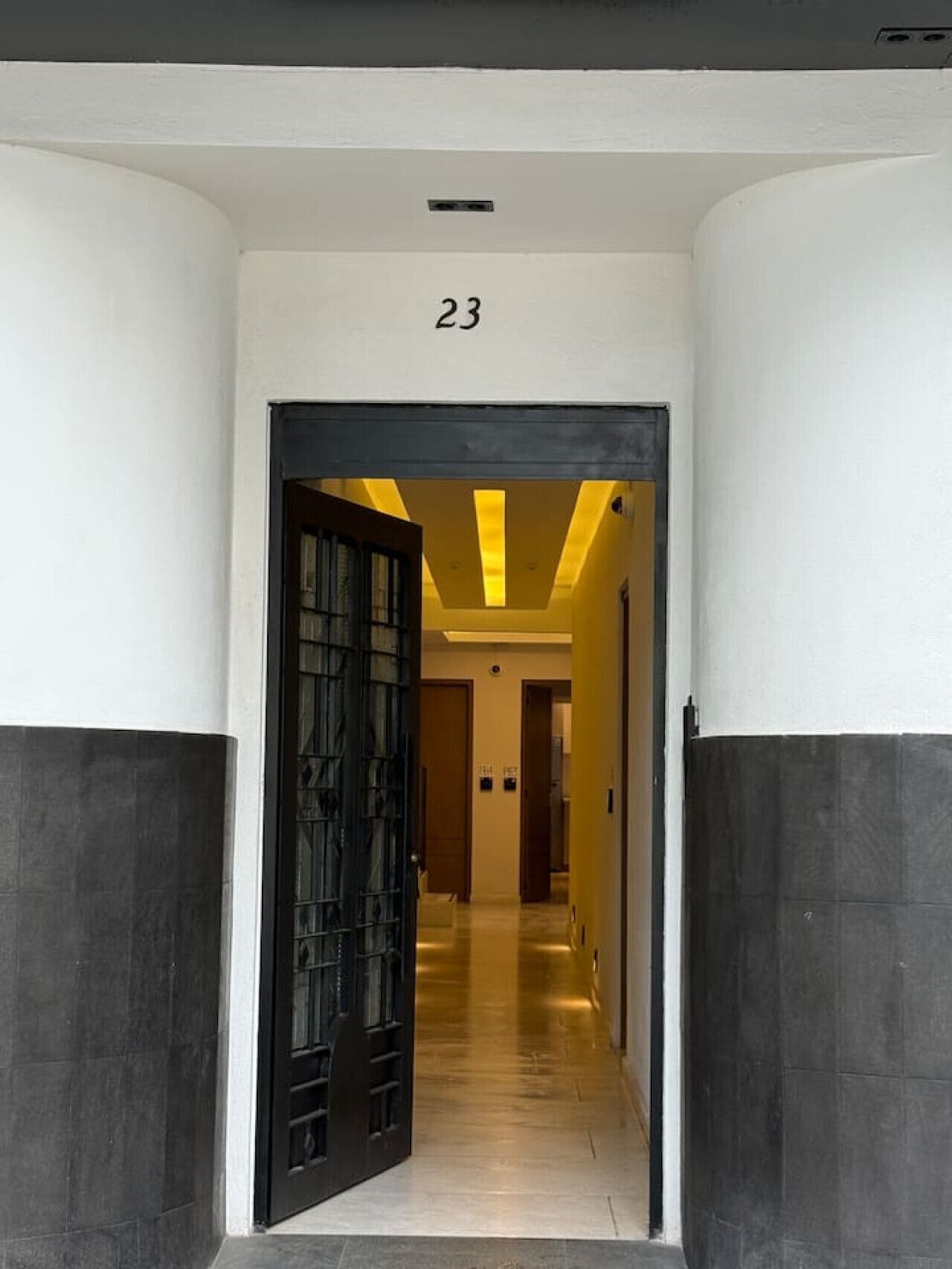 Un pasillo con una puerta negra marcada con el número 23, que da a una habitación con piso de baldosas e iluminación empotrada en el techo.