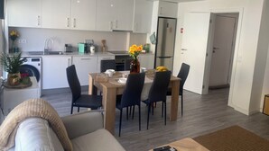 Moderni keittiö, jossa on valkoiset kaapit, jääkaappi sekä ruokailutila, jossa on pöytä ja tuolit.