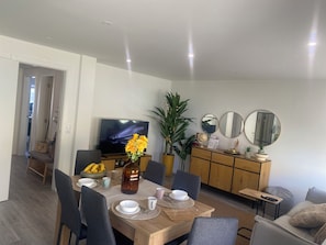 Ein moderner Essbereich mit einem Holztisch, gedeckt für vier Personen, umgeben von grauen Stühlen. Gegenüber vom Tisch ist ein Fernseher an der Wand befestigt.
