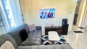 Un salon avec un canapé d’angle gris, une table basse, un téléviseur à écran plat fixé au mur et un tapis à motifs sur le sol.