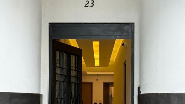 Ein Flur mit einer schwarzen Tür und der Zahl 23 darüber, der zu einem Raum mit gelber Decke und einer Holztür führt.