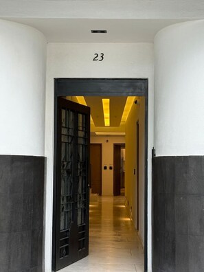 En hall med en svart dörr och siffran 23 ovanför, som leder in till ett rum med ett gult tak och en trädörr.