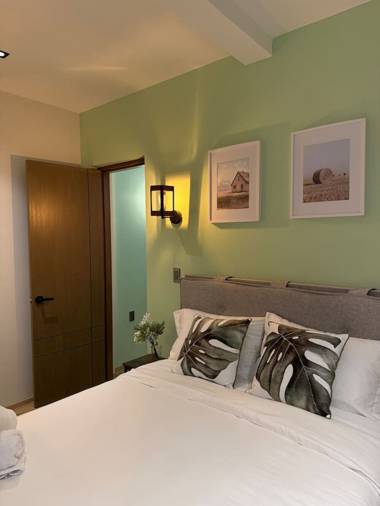 A bedroom with a bed, a nightstand, a wall-mounted lamp, and two framed pictures.