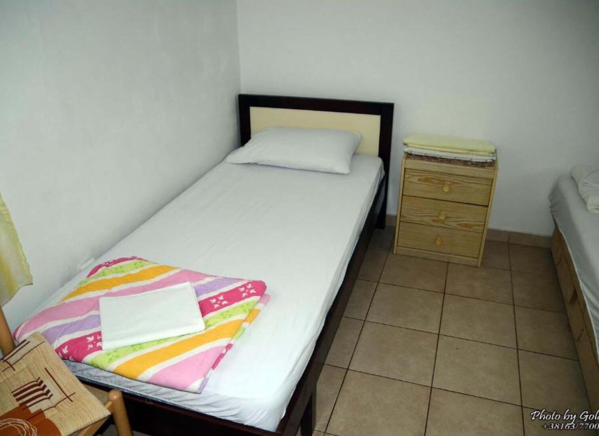 A small, neatly arranged bedroom with a single bed, a wooden nightstand with drawers, and a small table with a lamp.