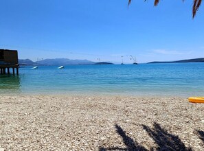Una playa de guijarros con agua cristalina, barcos y montañas al fondo.