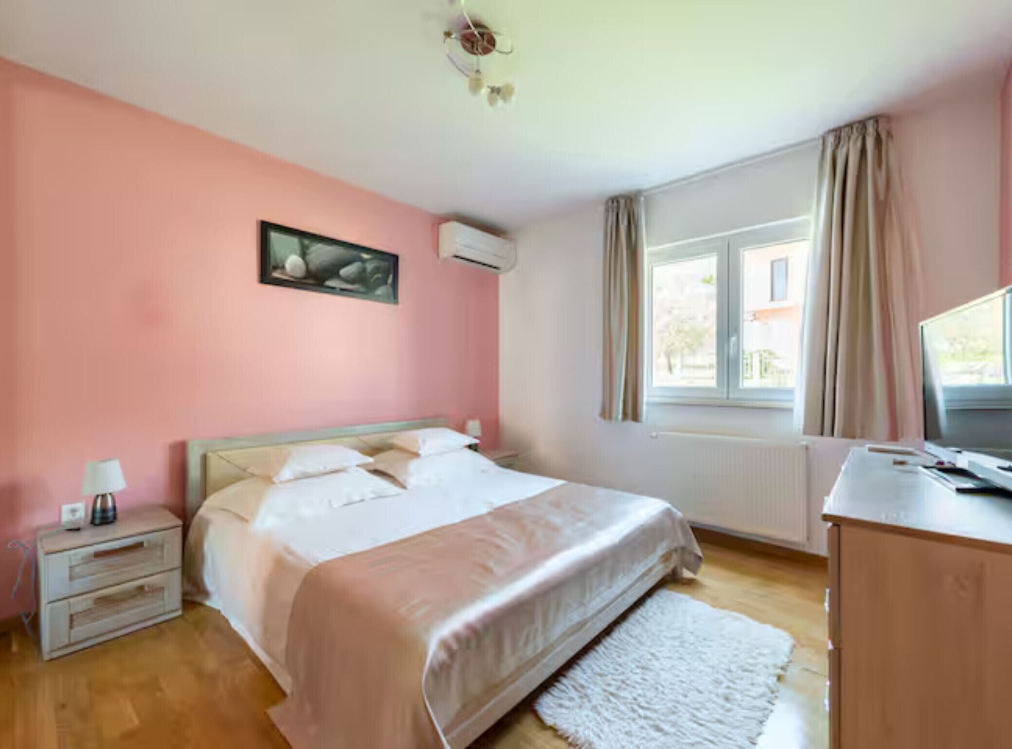 A bedroom with a bed, a nightstand, a desk, a window with curtains, and an air conditioning unit.