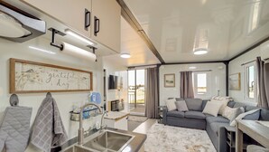 A modern kitchen with a stainless steel sink, a microwave, and a kettle. There is a gray quilted oven mitt hanging on the wall, and a framed quote on the wall.