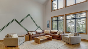 A modern living room with a sectional sofa, a coffee table, and large windows offering a view of trees. - Luxe Mountain Home w/ Chef’s Kitchen & Okemo Views (Ludlow)