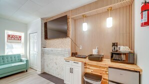 Cozinha compacta com micro-ondas, pia e pequena geladeira. Possui bancada de madeira e armário branco. Um sofá verde é visível na área de estar adjacente.