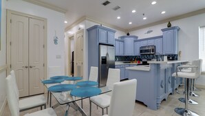Een moderne keuken met blauwe kastjes, een centraal eiland met barkrukken, en een eethoek met een glazen tafel en witte stoelen.