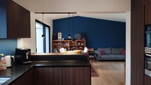 Eine moderne Küche mit dunklen Holzschränken, einer schwarzen Arbeitsplatte und einem Essbereich mit Holztisch und Stühlen. Im Wohnbereich befindet sich ein Bücherregal mit verschiedenen Gegenständen sowie ein graues Sofa.