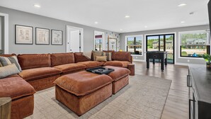 A spacious living room with a large sectional sofa, a coffee table, and a view of the outdoors through large windows. - *NEW*Private Chef-Family Friendly-Beach-Arcade Games-WI Dells (New Lisbon)