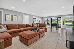 A spacious living room with a large sectional sofa, a coffee table, and a view of the outdoors through large windows. - *NEW*Private Chef-Family Friendly-Beach-Arcade Games-WI Dells (New Lisbon)