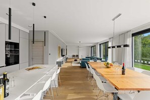 Eine moderne Küche mit einem langen Esstisch, weißen Stühlen und einem Holztisch mit einer Weinflasche und Obst.