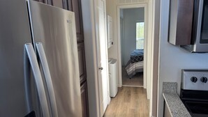 Et aflangt køkken med et køleskab, ovn og mikroovn. En dør fører til et soveværelse med seng og vindue.