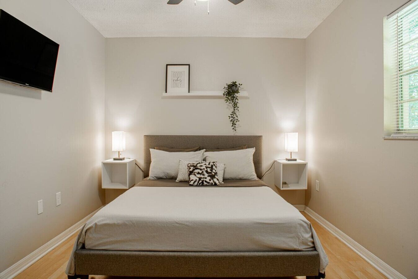 A bedroom with a bed, two bedside tables, a wall-mounted TV, a framed quote, and a window with blinds.