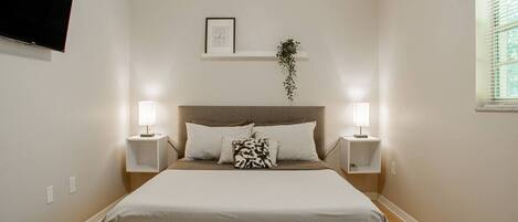 A bedroom with a bed, two bedside tables, a wall-mounted TV, a framed quote, and a window with blinds.