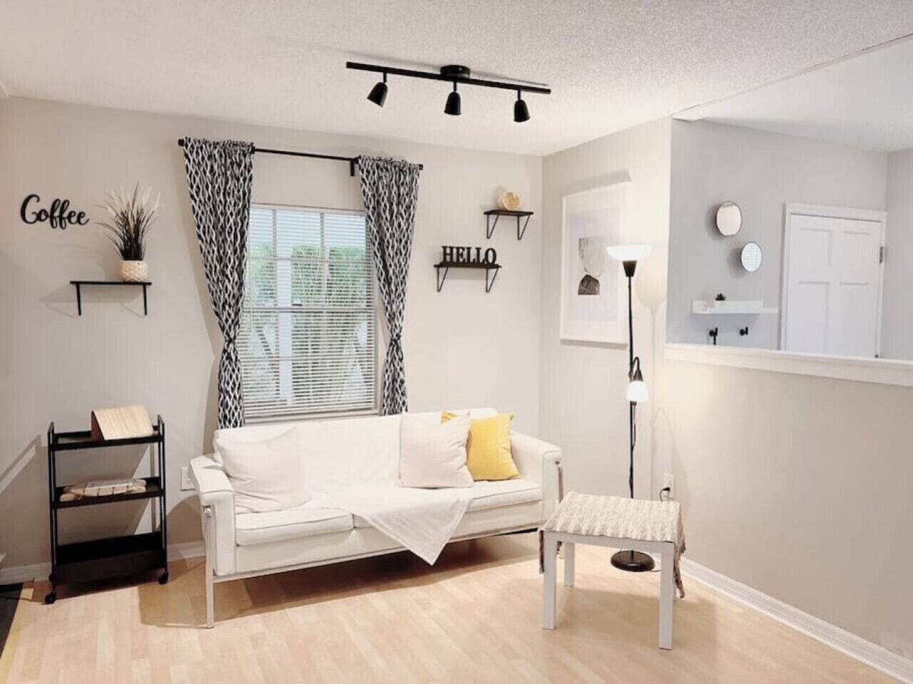 A modern living room with a white sofa, a bench, a coffee-themed decor, and a window with patterned curtains.
