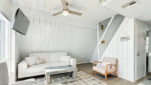 A modern living room with a white sofa, a wooden coffee table, and a light-colored armchair. A ceiling fan and a mounted TV are visible. - Iconic A Frame Directly Over The Surfside Waves (Surfside Beach)