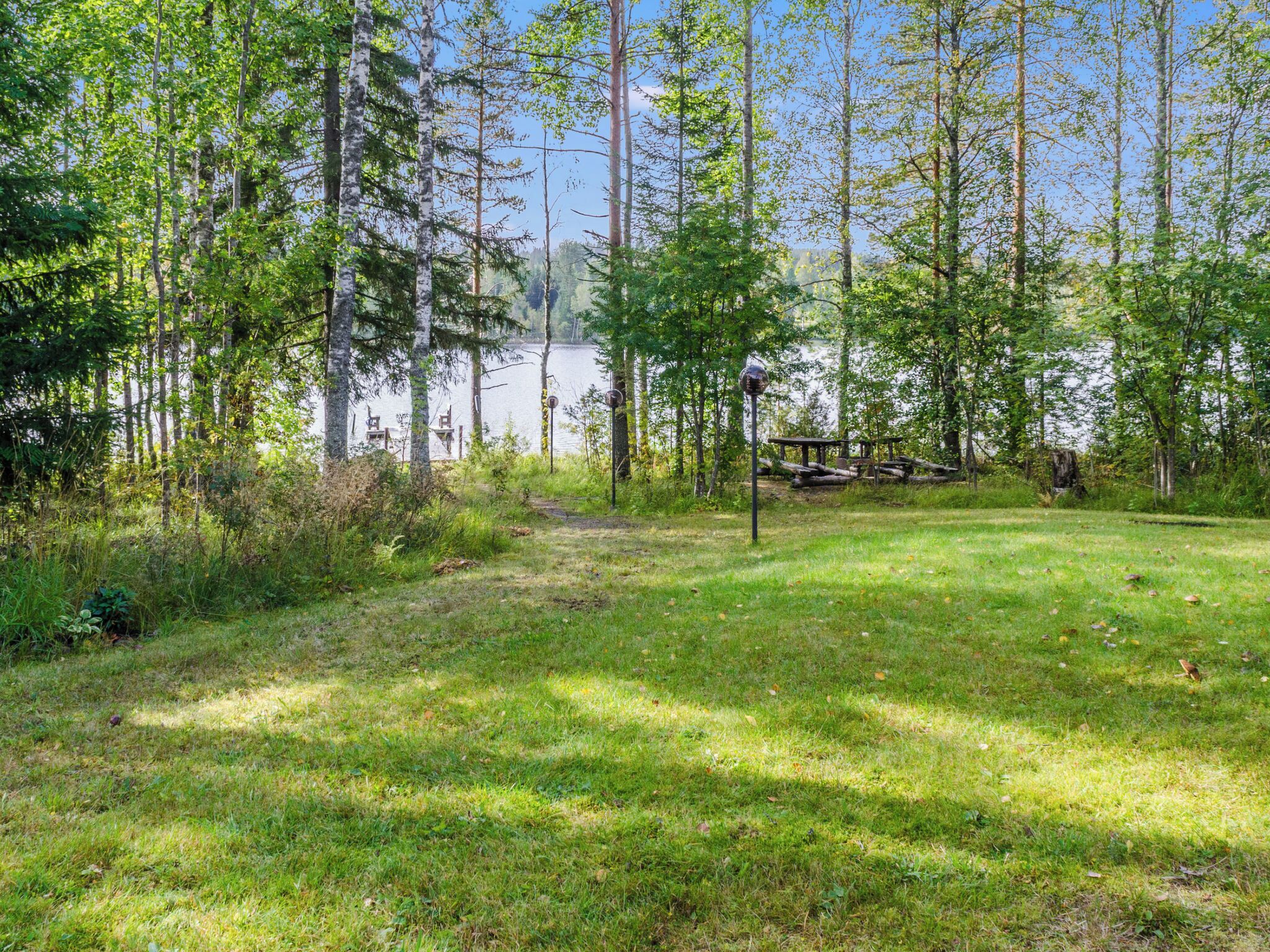 Une zone herbeuse avec un banc en bois et un lampadaire en métal, entourée d’arbres et bordée par une étendue d’eau.