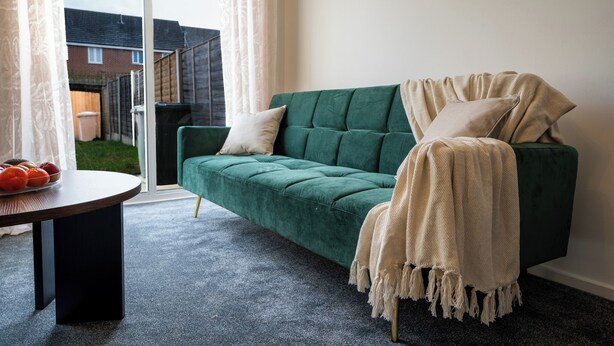 Un divano verde con una coperta beige, un tavolino con della frutta e vista su un giardino tramite una porta di vetro.