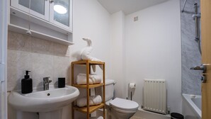 Un bagno con lavandino, water e doccia bianchi. Si nota una mensola in legno con asciugamani e un cestino nero.