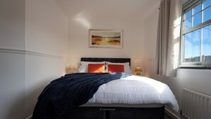 Un letto rifatto con copriletto bianco e blu, due cuscini e una coperta in blu scuro. Sopra il letto, un quadro incorniciato raffigura un paesaggio, e a destra si trova una finestra con tende.