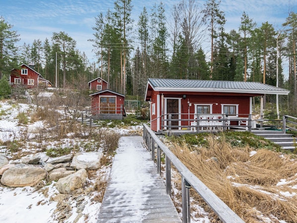 A wooden pathway leading to a red cabin with a balcony, surrounded by pine trees and snow.