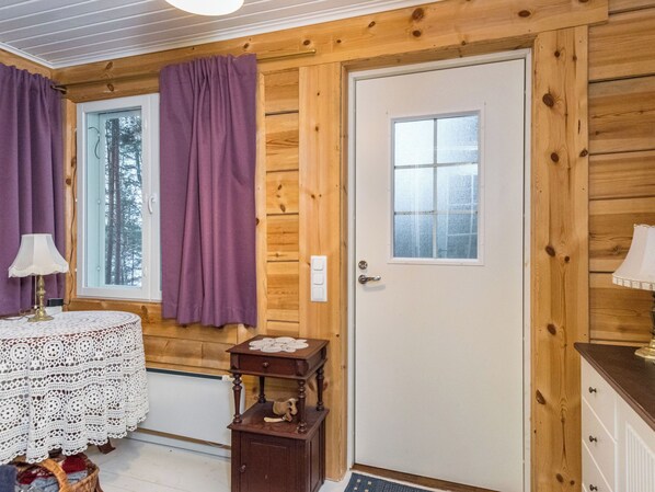 A wooden cabin interior with a door, a window, a small table with a lace tablecloth, and purple curtains.