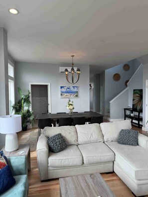A living room with a beige sectional sofa, a wooden coffee table, a dining table with chairs, a chandelier, and a staircase. - Beautiful Beach Views at The Sea View Estate! (Long Beach)