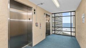 A hallway with two elevators, a fire alarm, and a large window with a view of the ocean. - Oceanfront, Pools, Lazy River @ Beach Cove Resort (North Myrtle Beach)