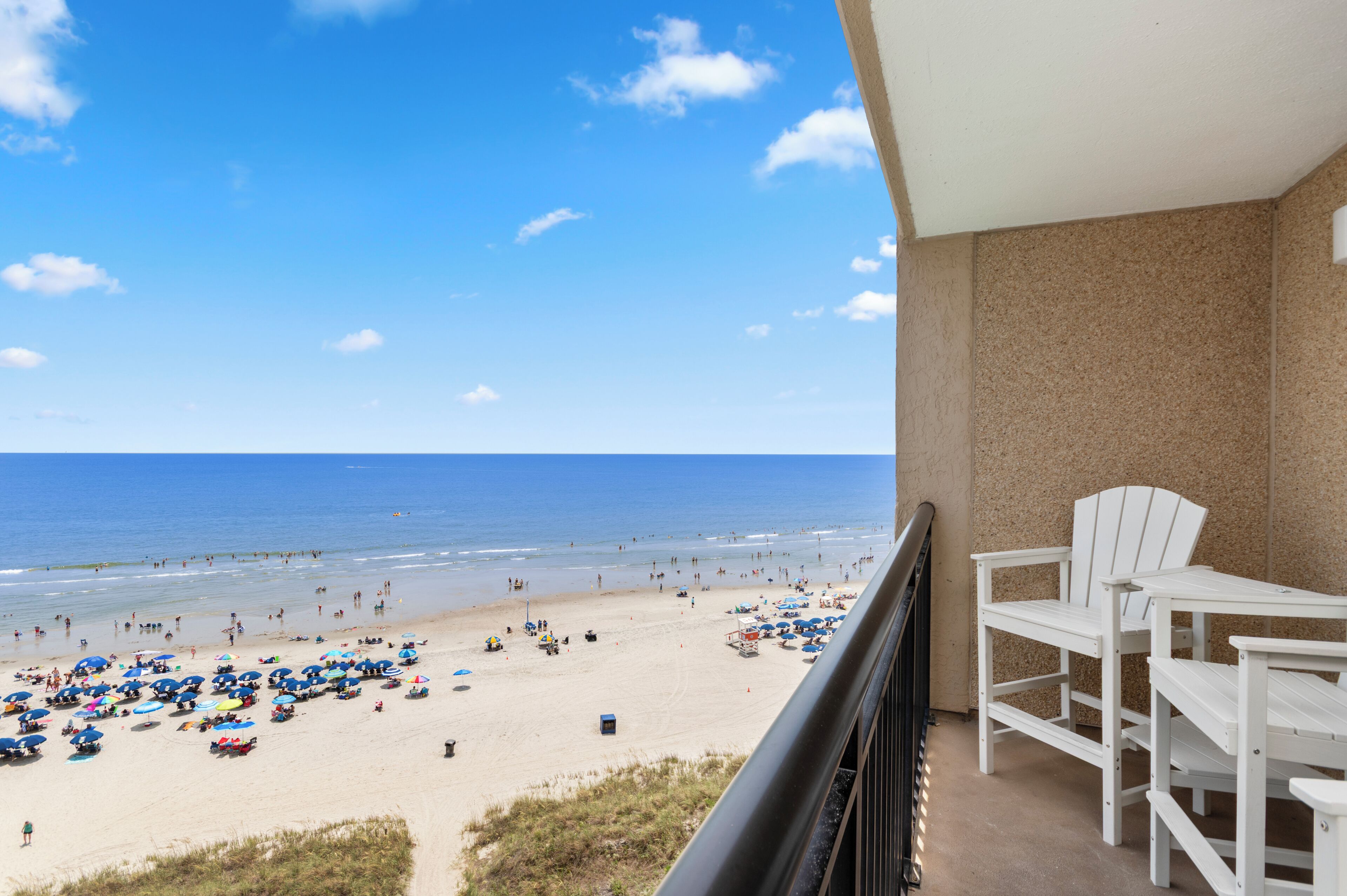 A balcony with a view of a beach, white chairs, and a table.