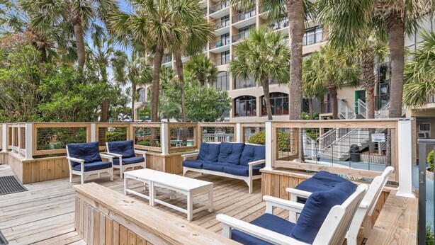 A wooden deck with outdoor seating, palm trees, and a building with balconies in the background. - Oceanfront, Pools, Lazy River @ Beach Cove Resort (North Myrtle Beach)