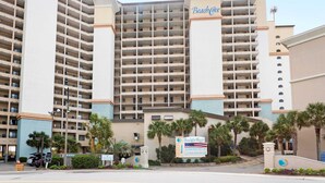 A large hotel with multiple floors, a prominent 'Beach Cove' sign, and a parking area with a palm tree-lined entrance. - Oceanfront, Pools, Lazy River @ Beach Cove Resort (North Myrtle Beach)