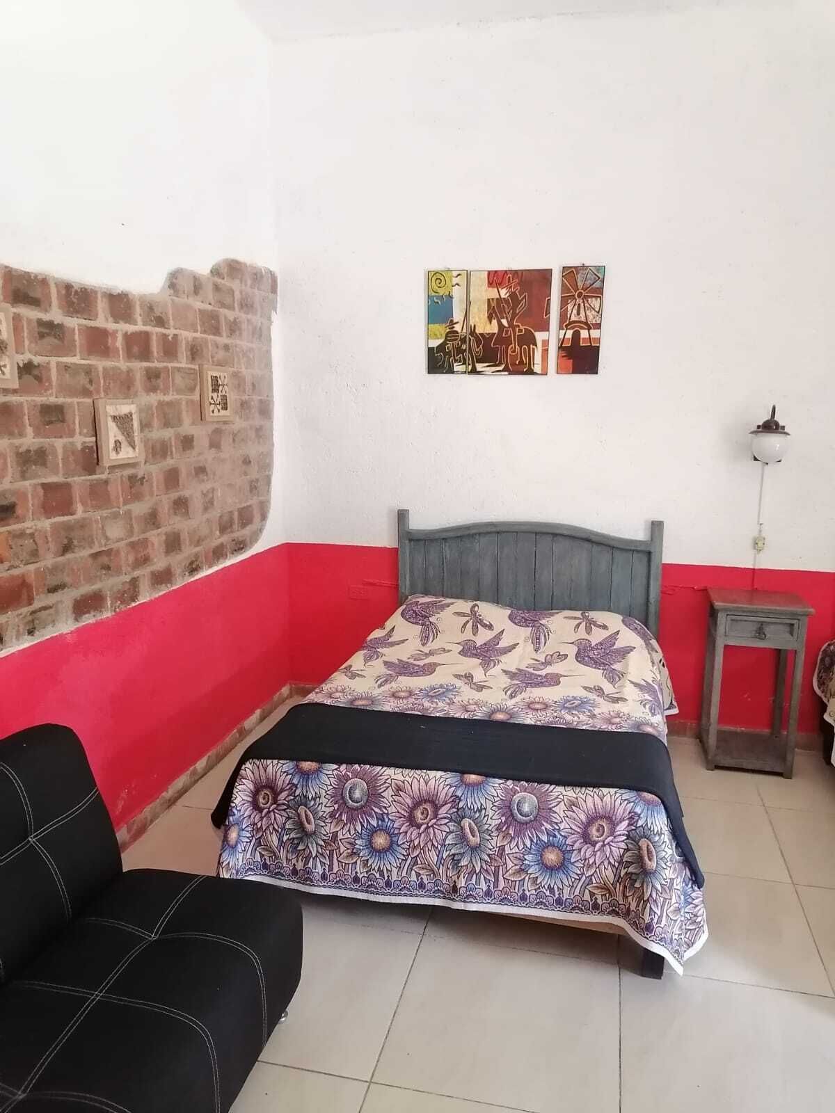 A bedroom with a bed, a brick wall, a red wall, a small table, and a wall-mounted lamp.