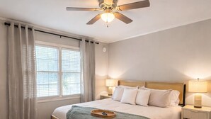 Un dormitorio con cama, lámparas de noche, ventilador de techo, ventana con cortinas y una alfombra estampada.