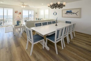 A dining area with a white table and chairs, a chandelier, and a view of the ocean through large windows. - Sea Salt - Surfside Beach (Surfside Beach)