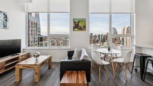A modern living room with a large TV, wooden furniture, a black sofa, white chairs, and a city view through large windows. - Stylish 1-Bedroom Apartment in Calgary | BLVD Beltline (Calgary)