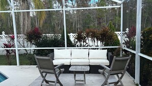 Un área de estar al aire libre techada con un sofá y dos sillones de mimbre, una mesita y vistas a una piscina y árboles.