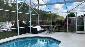 Una piscina exterior techada con estructura blanca, un sofá negro y dos sillas.
